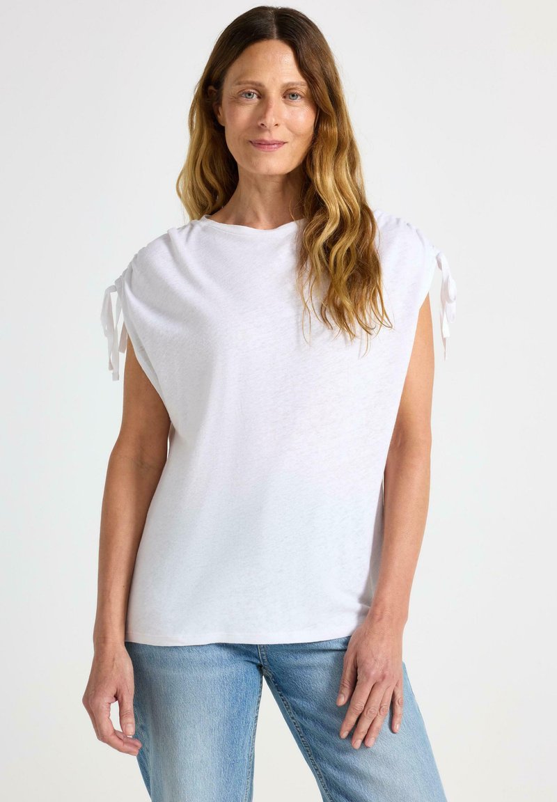 Hvid kortærmet top med samlede skuldre, lavet af et blødt stof. Har bundet detaljer på ærmerne og en afslappet pasform. Bæres med blå jeans.