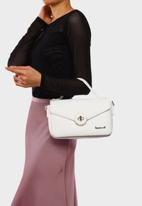Borsa a mano in pelle bianca con finitura texturizzata, chiusura a patta rotonda, hardware in argento e manico superiore, tenuta accanto a un modello con una maglietta nera.