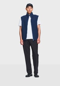 Jeune homme portant un gilet sans manches bleu marine sur un t-shirt blanc, pantalon noir, chaussures noires et une casquette noire, debout devant un fond clair uni.