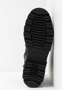 Semelle noire de botte UGG avec des motifs de bande de roulement profonds et larges pour la traction, positionnée verticalement contre un fond blanc à angles.