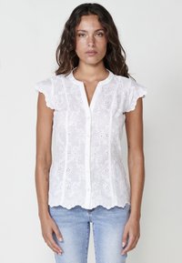 Blouse blanche brodée avec des manches en capes festonnées, un devant à boutons, des motifs floraux et un ourlet arrondi. Portée avec un pantalon en denim bleu.