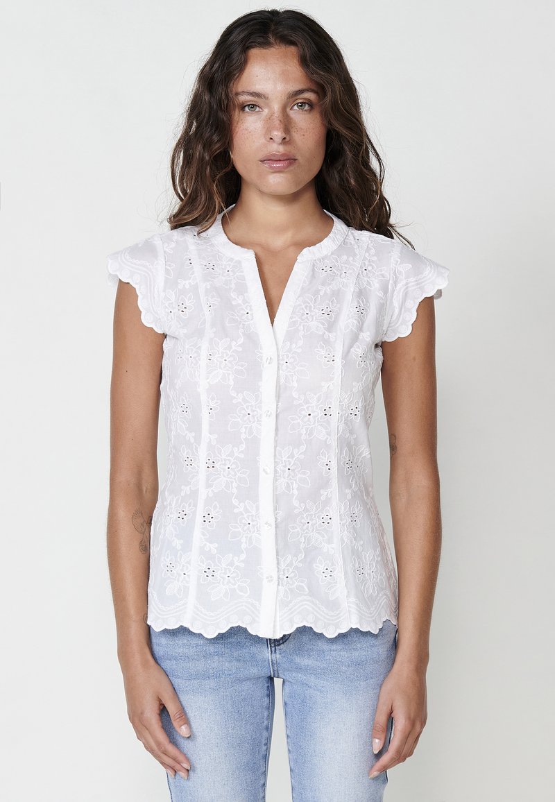 Blouse blanche brodée avec des manches en capes festonnées, un devant à boutons, des motifs floraux et un ourlet arrondi. Portée avec un pantalon en denim bleu.
