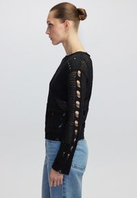 Pull en maille noire avec un design ajouré, manches longues avec des motifs découpés, et un col rond, associé à un jean bleu clair.