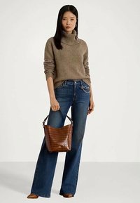 Žena nosi smeđi pulover s visokim ovratnikom i plave traperice, držeći smeđu kožnu torbu s lancem.