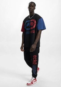 Black graphic t-shirt with red and blue sleeves, featuring a logo and text. Black sweatpants with "eckō unltd" by the leg, paired with red sneakers.