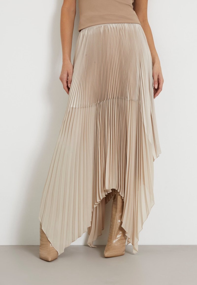 Jupe plissée beige asymétrique portée avec des bottes beige, vue de la taille aux pieds sur un fond clair uni.