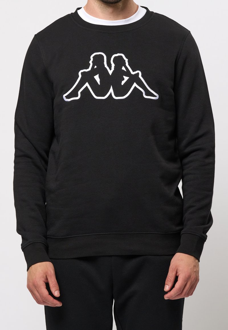 Homme portant un sweat-shirt noir avec un logo en silhouette blanche de deux personnes assises dos à dos sur la poitrine.
