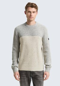 Junger Mann, der einen hellgrauen und cremefarbenen, farbblockierten Strickpullover und dunkelgraue Jeans trägt, vor einem schlichten Hintergrund steht.