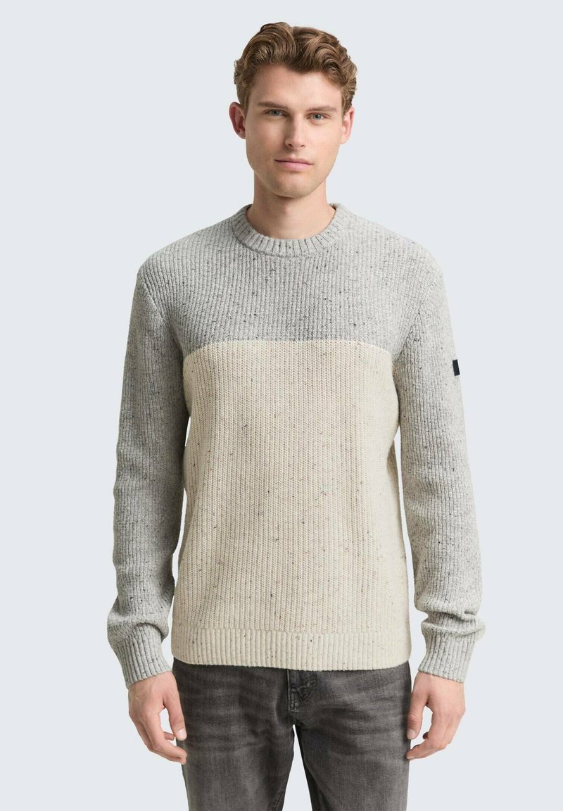 Junger Mann, der einen hellgrauen und cremefarbenen, farbblockierten Strickpullover und dunkelgraue Jeans trägt, vor einem schlichten Hintergrund steht.