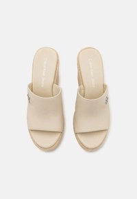 Sandalias deslizantes beige con parte superior de tela suave, punta abierta y suela de yute trenzado. Presenta un pequeño adorno en el lateral.
