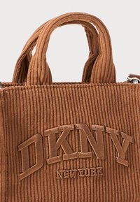 Brown corduroy handbag featuring double handles and embroidered "DKNY NEW YORK" logo. Textured surface with a boxy shape and metallic accents.