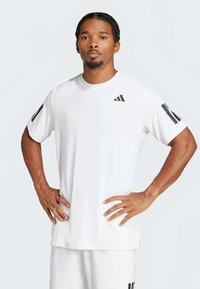 Bela atletska majica iz lahkega blaga, z rdečim logotipom Adidas in črnimi črtami na rokavih, z klasičnim okroglim izrezom.