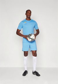 Heleroheline spordisärk kuldsete detailidega, sobivad lühikesed püksid, valged põlvikud ja mustad jalgpallikengad; hoiab käes musta-valget jalgpalli.