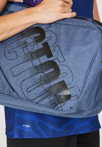 Blå tygaxelväska med svart grafisk textdesign. Inkluderar dragkedjestängning och texturerade inslag längs kanterna.