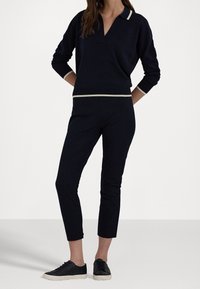 Nainen, jolla on yllään tummansininen pitkähihainen paita vaaleanbeigeillä yksityiskohdilla ja siihen matchaavat lyhyet housut, sekä mustat lenkkarit valkoisilla pohjilla.