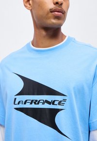 Νέος άνδρας που φοράει ένα ανοιχτό μπλε t-shirt LaFrance με μεγάλο μαύρο λογότυπο στο στήθος, κοιτάζοντας προς το πλάι σε απλό φόντο.