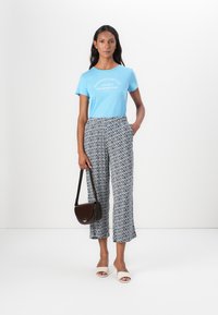 T-shirt azzurro chiaro con testo bianco, pantaloni a gamba larga neri e bianchi a fantasia, sandali beige e una piccola borsa a tracolla marrone.
