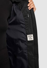 Zwarte jas met een binnenvoering van donker stof, voorzien van een ritstaskzak en een label met de tekst "Een warme omhelzing gemaakt van stof."