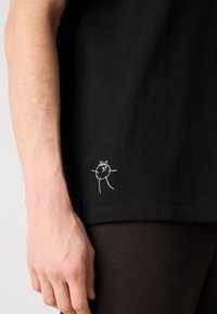Black cotton t-shirt featuring a hand-drawn white figure embroidered on the lower left side, showcasing a relaxed fit and smooth texture.