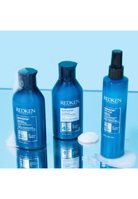 Tre Redken hårvårdsprodukter: blå flaskor med schampo, balsam och anti-snap-behandling med svarta lockar på en reflekterande yta.