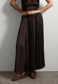 Bruine geplooide rok met elastische tailleband, gemaakt van een vloeiend materiaal, gecombineerd met bruine ballerina's, gepresenteerd tegen een neutrale achtergrond.