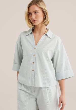 Femme portant une chemise à boutons à rayures verticales bleu clair et blanc avec des manches trois-quarts et un pantalon assorti, debout devant un fond uni.