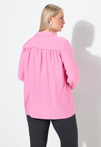 Hellrosa, leichtes Langarmshirt mit einem Rückenfalte, Button-Down-Kragen und weicher Textur, kombiniert mit dunkelgrauen Leggings.