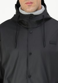 Veste noire étanche avec un col haut, un col côtelé gris, des boutons à l'avant et une capuche avec cordon de serrage. Comprend un patch logo.