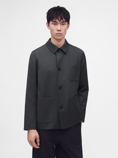 Jeune homme aux cheveux foncés portant une veste grise foncée boutonnée avec trois poches, debout devant un fond blanc uni.