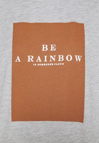 Grå t-shirt med ett rostfärgat rektangeldesign och vit text: "VAR EN REGNBÅGE I NÅGONS MOLN." Mjuk textur, avslappnad stil.