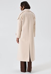 Lungo cappotto beige realizzato in tessuto liscio, dotato di una fessura posteriore, spalline e polsini con bottoni, indossato sopra pantaloni marroni.