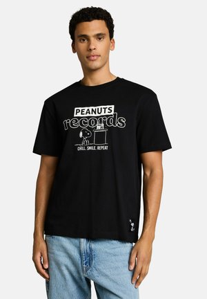 Junger Mann trägt ein schwarzes T-Shirt mit weißem "Peanuts records"-Text und Snoopy-Grafik, kombiniert mit hellblauen Jeans, steht vor weißem Hintergrund.