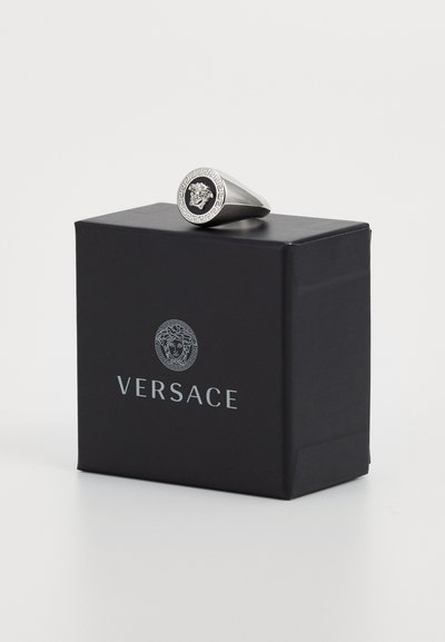 Sudraba gredzens ar apaļu melnu centru, kam ir izvirzīts raksts, novietots uz kastes, kurā izdrukāts Versace logotips baltā krāsā un Grieķu atslēgas raksts.