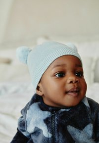 Ljusblå ribbad stickad mössa med pom-poms; marinblå fleeceoutfit med stjärnmönster. Nere bild på en babys ansikte och uttrycksfulla ögon.