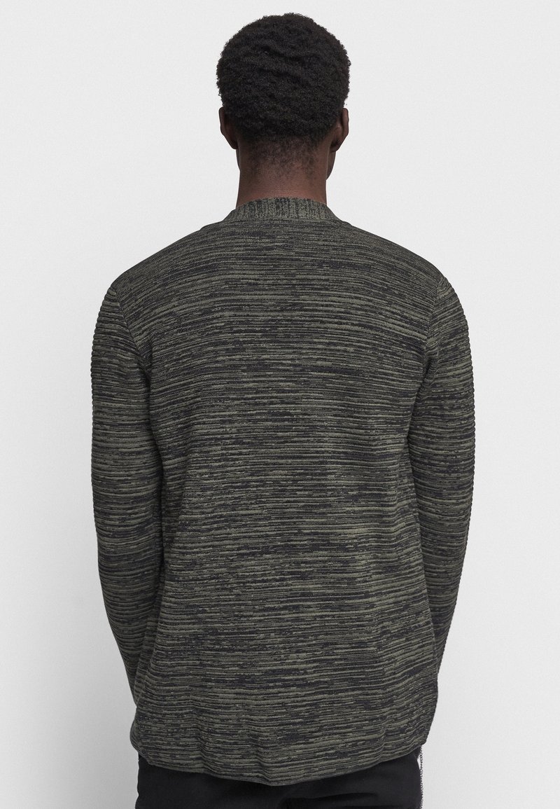 Homme portant un pull texturé à rayures gris foncé et noir, vu de dos sur un fond blanc uni.