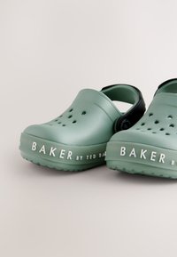 Groene rubberen klompen met ventilatiegaten, zwarte accenten en een wit logo met de tekst "BAKER by TED BAKER" op de zool. Textuurafwerking.