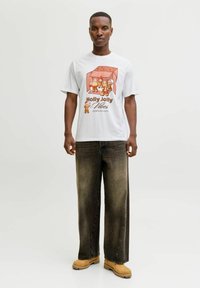 Bărbat în picioare, purtând un tricou alb cu figurine de turtă dulce și textul "Holly Jolly Vibes", blugi întunecati largi și cizme bej, pe un fundal alb.