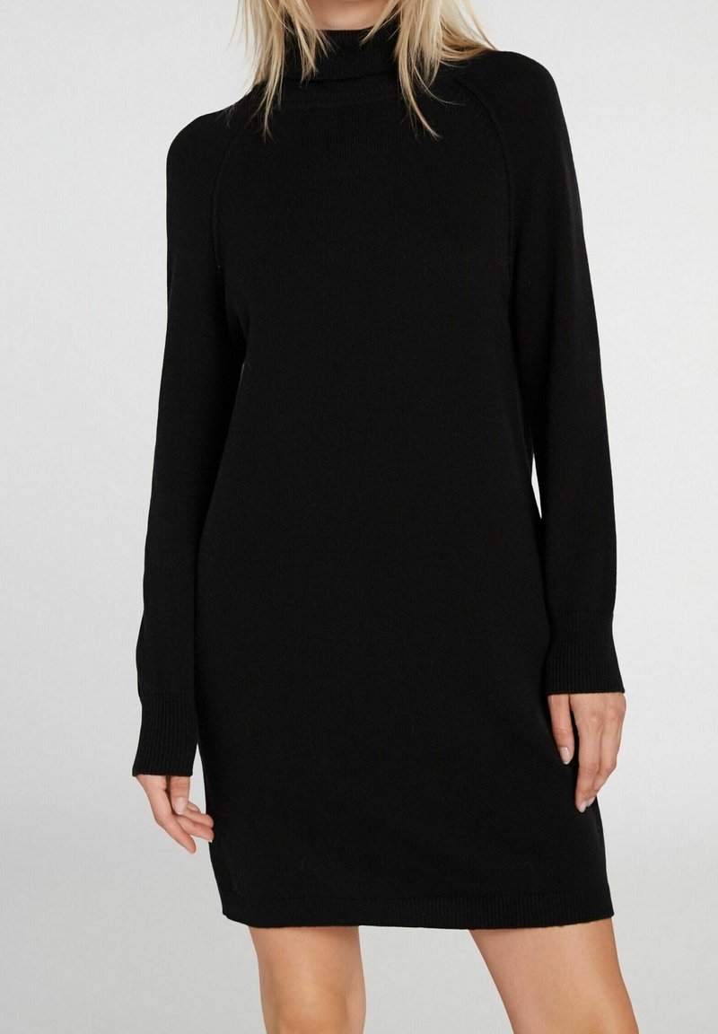 Femme portant une robe en maille noire, à manches longues, longueur genou, avec un col montant, posant devant un fond clair uni.