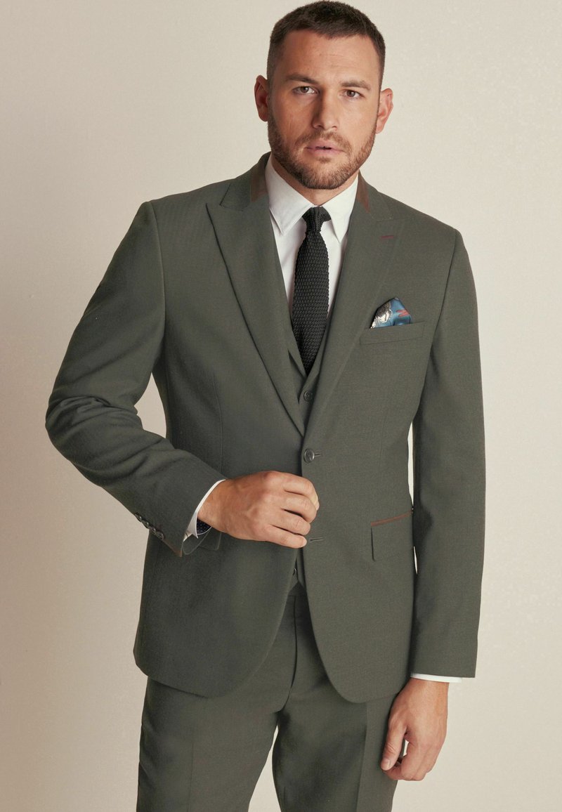 Dark green suit with a textured finish, slim fit, single-breasted design, white shirt, and black tie. Pocket square features blue and red patterns.