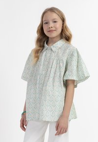 Camicia a maniche corte di colore verde chiaro con un motivo a foglie, dotata di un colletto classico e dettagli arricciati. Indossata con pantaloni bianchi.