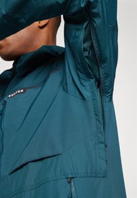 Veste teal en matière légère et résistante à la déchirure. Comprend des aérations doublées en mesh, des zips visibles et un logo sur la zone de la poitrine.