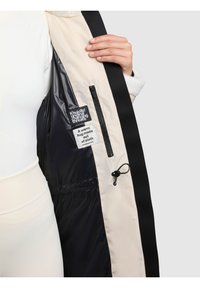 Svart och beige vadderad jackas insida med en glänsande textur, utrustad med en dragkedjeficka, etiketter och en dragsko för justering.