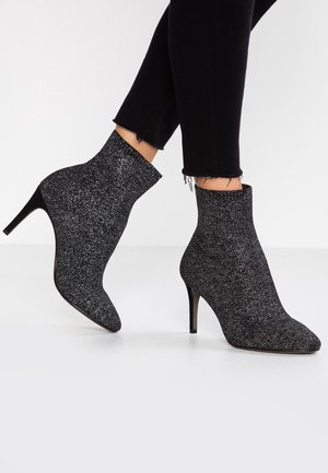 Woman wearing black and silver glitter ankle boots with pointed toes and stiletto heels, paired with black cropped pants.