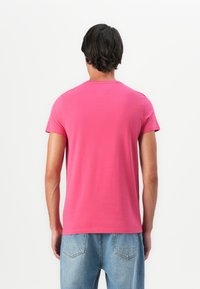 Un homme se tenant de dos, portant un t-shirt rose vif à manches courtes et un jean en denim bleu clair, contre un fond blanc uni.