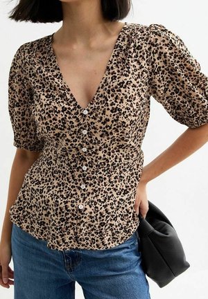 Kvinde iført en beige leopardmønstret skjorte med knapper og pufærmer samt blå jeans, der holder en sort tekstureret håndtaske.