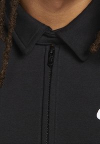 Svart zip-up jacka med krage. Har en hel dragkedja och en vit logotyp på vänster sida. Slät textur, avslappnad design.