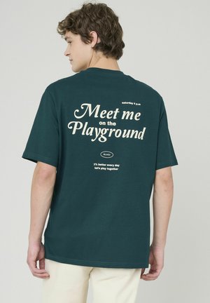 T-shirt en coton teal avec une coupe décontractée, arborant du texte blanc au dos : "Retrouve-moi sur le terrain de jeu," et d'autres phrases ludiques en dessous.