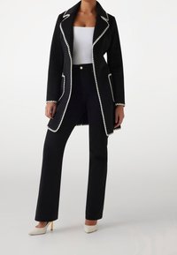 Abrigo negro con detalles de costura blanca, mangas largas y cuello con muesca, combinado con pantalones negros y tacones blancos de punta.