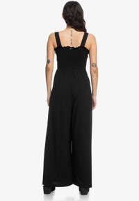 Kvinna med långt hår i en svart ärmlös jumpsuit, stående med ryggen mot kameran, visar tre tatueringar på ryggen och armarna.