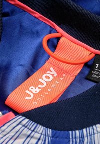 Mörkblå ytterklädestyg med en texturerad krage; har en livlig orange etikett med vit text "J&JOY OUTERWEAR" och en cirkulär accent.